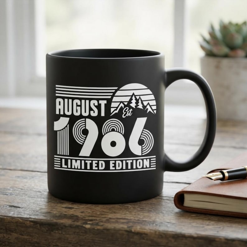 May include: A matte black ceramic mug featuring white printed text and graphics. The design includes the words AUGUST EST 1986 LIMITED EDITION, accompanied by a stylized mountain range and sun graphic with horizontal line accents.