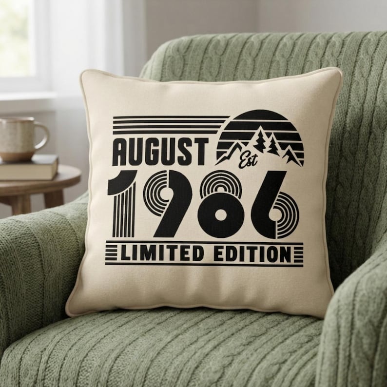 May include: A square cream-colored throw pillow featuring black graphic text and imagery. The design includes the words AUGUST, EST, and LIMITED EDITION, along with the year 1986 in large stylized font and a mountain sunset graphic.