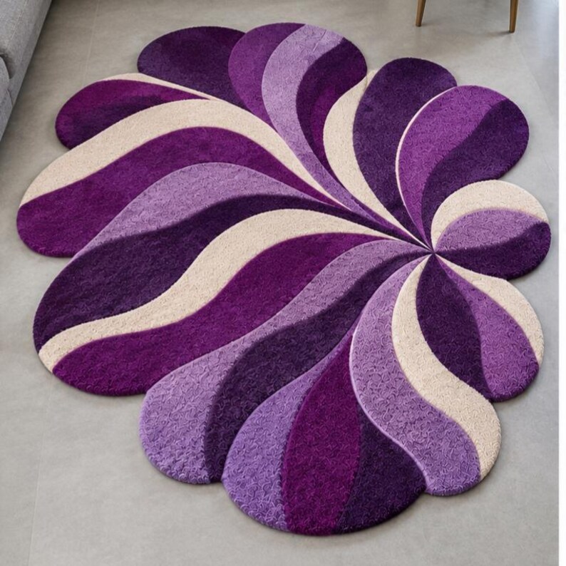 May include: A circular rug featuring a repeating pattern of curved, petal-like shapes in shades of deep purple, light violet, and cream. The textured surface consists of dense, tufted fibers arranged in a swirling, fan-like radial design.