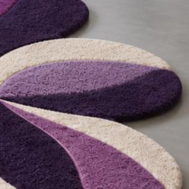 May include: A rug featuring a floral-inspired shape with rounded petals. The surface is composed of textured, swirling patterns in alternating shades of deep purple, light lavender, and cream, creating a layered and dimensional appearance.