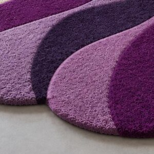May include: A textured rug featuring a series of overlapping, curved segments in varying shades of purple, ranging from deep violet to lighter lavender. The surface has a dense, looped pile construction that creates a consistent, repeating pattern.