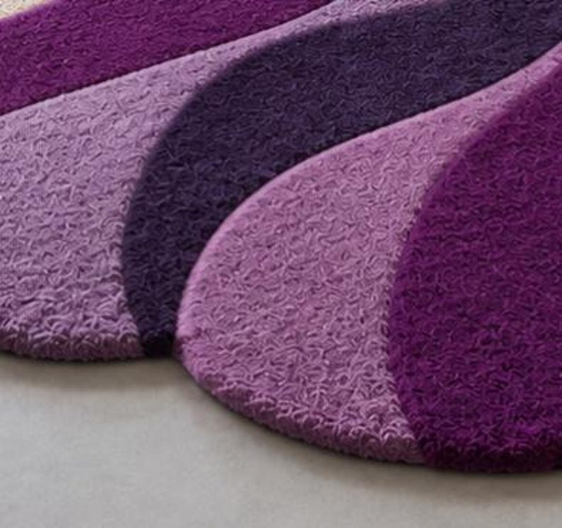 May include: A textured rug featuring a series of overlapping, curved segments in varying shades of purple, ranging from deep violet to lighter lavender. The surface has a dense, looped pile construction that creates a consistent, repeating pattern.