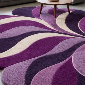 May include: A large, plush area rug featuring a repeating, wave-like pattern in shades of deep purple, lavender, and cream. The rug has a thick, textured pile and a curved, organic shape that extends across the floor, creating a layered visual effect.