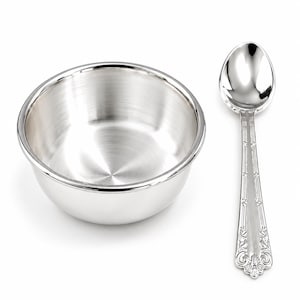 Op de afbeelding: Een gepolijste, zilverkleurige kom met een gladde, ronde binnenkant en een bijpassende zilverkleurige lepel met een sierlijk, in reliëf aangebracht bloemmotief op het handvat. Beide items hebben een reflecterende afwerking en een strak ontwerp.