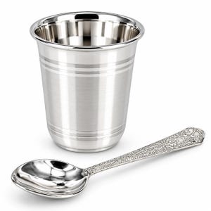 Puede incluir: Un vaso de metal plateado pulido con bandas horizontales alrededor de la parte superior e inferior, junto con una cuchara de metal plateado a juego que presenta un intrincado patrón floral y geométrico en relieve en el mango y una cazoleta lisa.