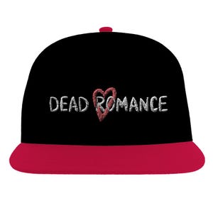 May include: A black baseball cap with a flat, bright pink brim and a top button. The front features the embroidered white text "DEAD ROMANCE" with a textured red heart replacing the letter "O" in the word "ROMANCE" for a bold, graphic look.