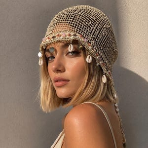 May include: A beige crochet headpiece featuring a wide-mesh net design adorned with a decorative trim of small pink beads, metallic sequins, and white cowrie shells that hang along the front and side edges of the cap in a fringe pattern.