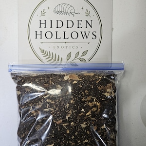 May include: A clear plastic resealable bag filled with dark brown organic substrate containing wood chips and soil. Behind the bag is a white rectangular card featuring a logo with a pill bug illustration and the text Hidden Hollows Exotics.