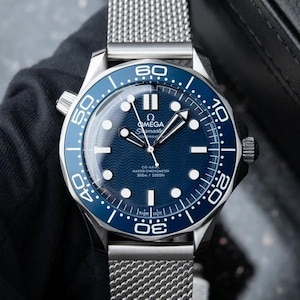 Op de afbeelding: Een roestvrijstalen Omega Seamaster horloge met een blauwe wijzerplaat met golfpatroon, lichtgevende uurmarkeringen en een draaibare ring met witte cijfers. Het horloge heeft een zilverkleurige mesh-band en toont de tekst Omega Seamaster Co-Axial Master Chronometer 300m / 1000ft op de wijzerplaat.