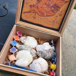 Könnte beinhalten: Eine Holzkiste mit offenem Deckel, der eine gedruckte Schatzkarte zeigt. Im Inneren befinden sich verschiedene natürliche Muscheln, ein Set kleiner bunter Farbtöpfe aus Kunststoff und ein dünner blauer Pinsel auf braunem Füllmaterial.