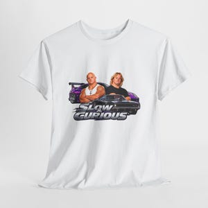 Op de afbeelding: Een wit T-shirt met korte mouwen en een grafische print van twee personen voor een paarse sportwagen en een zwarte muscle car. Onder de figuren staat de tekst SLOW & CURIOUS in een vetgedrukt, metaalachtig lettertype.
