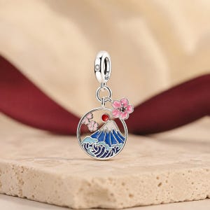 Op de afbeelding: Een zilveren bedel met een cirkelvormig frame met een blauw-witte berg, een rode zon en roze kersenbloesems. De bedel heeft een klein oogje met S925-stempel en een heldere steen, met een extra roze bloemaccent dat eraan vastzit.