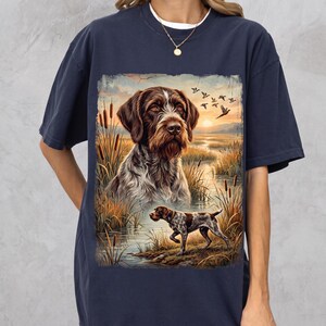 May include: A navy blue short-sleeve t-shirt featuring a graphic print of a German Wirehaired Pointer dog in a marshland setting with cattails, a sunset, and flying birds. The design includes a large portrait of the dog and a smaller full-body view.