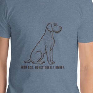 May include: A heather blue short-sleeve t-shirt featuring a dark brown line art illustration of a sitting dog in profile. Below the dog, the text reads: GOOD DOG. QUESTIONABLE OWNER. The shirt has a crew neckline and a textured fabric appearance.