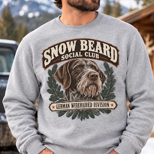 May include: A heather gray crewneck sweatshirt featuring a graphic print of a German Wirehaired Pointer dog. The text reads SNOW BEARD SOCIAL CLUB and GERMAN WIREHAIRED DIVISION, framed by dark green pine branches and two small red stars.