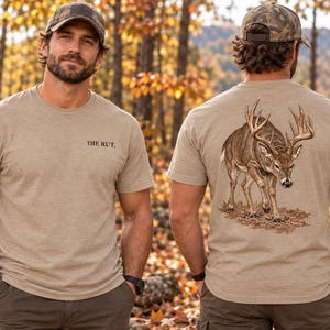May include: A tan short-sleeved t-shirt featuring the text THE RUT. printed in black on the front left chest. The back of the shirt displays a detailed graphic illustration of a large buck deer standing on brown leaves and forest ground.