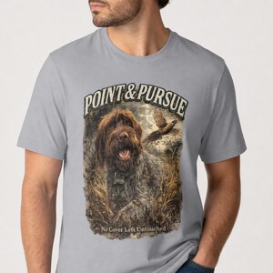 May include: A light gray t-shirt featuring a graphic print of a brown wirehaired pointing dog in tall grass with a flying pheasant. The text reads POINT & PURSUE at the top and No Cover Left Untouched at the bottom in a dark, weathered font.