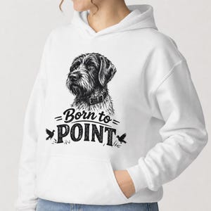 Op de afbeelding: Een witte hoodie met een zwarte grafische print van een draadharige staande hond. Onder de hond staat de tekst Born to POINT in een vet, gestileerd lettertype, geflankeerd door twee kleine vogel-silhouetten aan weerszijden van het woord.