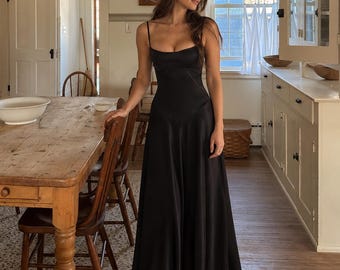 May include: A floor-length black satin dress featuring a scoop neckline, thin spaghetti straps, and a fitted bodice that transitions into a flowing, full-length skirt. The garment has a smooth, reflective finish and a structured waistline seam.