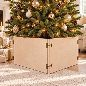 Puede incluir: Un cubrepies para árbol de Navidad de madera clara con forma cuadrada, que cuenta con bisagras metálicas y dos cierres de gancho en tono bronce en el panel frontal. El cubrepies rodea la base de un árbol decorado con adornos dorados.