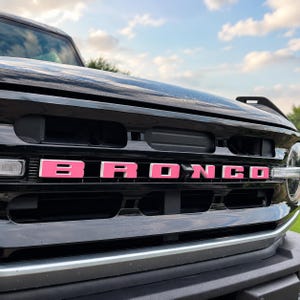 May include: The front grille of a black vehicle featuring the word BRONCO in bright pink lettering. The grille has a horizontal design with black slats and a silver trim piece along the bottom edge, set against a sky with clouds in the background.