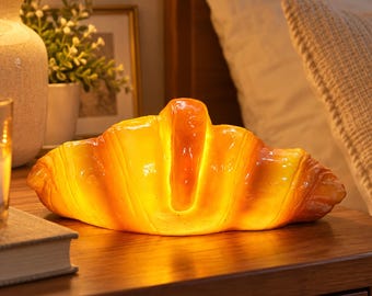 May include: A croissant-shaped lamp with a glossy, golden-brown finish that emits a warm, soft glow from within. The light fixture features realistic pastry folds and textures, resting horizontally on a flat surface to provide ambient illumination.