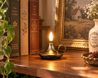 May include: A black and gold metallic oil lamp with a circular handle and a single glowing flame-shaped bulb. The lamp sits on a wooden surface next to three vintage books titled The Secret Garden, A World of Flowers, and The Art of Home.