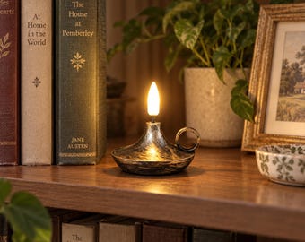 May include: A metallic oil lamp with a distressed silver and black finish, featuring a circular base, a small side handle, and a single glowing white flame-shaped bulb. The lamp sits on a wooden shelf next to several hardcover books.