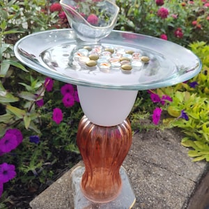 May include: A decorative bird bath constructed from stacked glass elements. It features a clear glass plate top filled with small, multicolored glass pebbles, a white frosted glass center section, and a ribbed amber glass base on a clear stand.