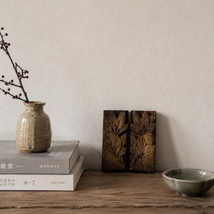 Könnte beinhalten: Eine beige Keramikvase mit einem dünnen Zweig, zwei gestapelte graue Bücher, zwei rechteckige Holzpaneele mit geschnitzten Blattmustern und eine kleine graue Keramikschale. Die Bücher zeigen chinesischen Text auf ihren Buchrücken.