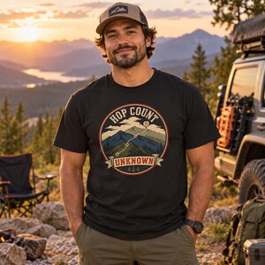 May include: A black short-sleeved t-shirt featuring a circular graphic print of a mountain landscape with a dotted trail path, the text "HOP COUNT" at the top, and the word "UNKNOWN" on a banner at the bottom, paired with olive green trousers.