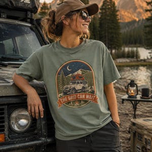 Op de afbeelding: Een saliegroen t-shirt met korte mouwen en een ronde grafische print van een terreinwagen met een daktent, dennenbomen, bergen en een maansikkel. De tekst THE GRID CAN WAIT staat op een banner onderaan de grafische afbeelding.