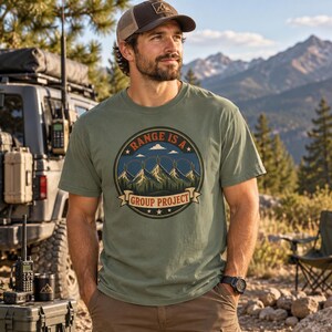 May include: An olive green short-sleeve t-shirt featuring a circular graphic on the chest. The design depicts a mountain range under a blue sky with the text "RANGE IS A GROUP PROJECT" printed in a circular border and on a banner at the bottom.