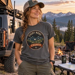 May include: A charcoal grey short-sleeve t-shirt featuring a circular graphic with a mountain range, a connected node network, and the text "EVERY NODE COUNTS" above a compass rose. The design uses white, yellow, and dark red colours on the fabric.