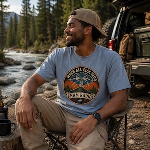 May include: A light blue t-shirt featuring a circular graphic with a radio tower, lightning, and the text "WHEN ALL ELSE FAILS HAM RADIO". The design includes a mountain landscape and a compass rose element at the bottom, printed on the chest.