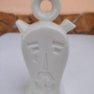 Può includere: Un contenitore in ceramica bianca con finitura lucida, caratterizzato da un volto stilizzato con occhi geometrici, naso triangolare e bocca frastagliata. L'oggetto presenta un manico circolare sulla parte superiore e due piccoli beccucci.