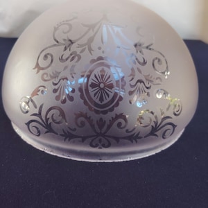 Op de afbeelding: Een koepelvormige lampenkap van matglas met een helder, geëtst decoratief patroon. Het ontwerp bestaat uit symmetrische krullen, bloemmotieven en een centraal ovaal medaillon met een ster-element, wat een contrast creëert tussen het matte oppervlak en de transparante glasdetails.