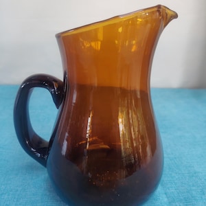 Op de afbeelding: Een doorschijnende, amberkleurige glazen kan met een rond lichaam, een gebogen handvat aan de zijkant en een kleine schenktuit aan de bovenrand. Het glas heeft een gladde, gepolijste afwerking en een diepe, warme bruine tint.