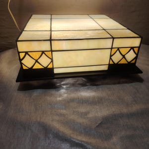May include: A square stained glass lamp with a flat top and sides featuring cream-colored panels. The side panels include a decorative lattice pattern in a darker amber hue, all held together by dark metal framing. A thin power cord extends from the top.