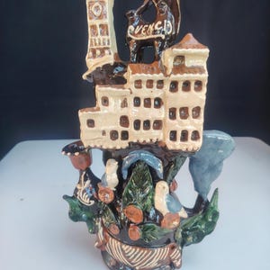 Może przedstawiać: Ceramiczna rzeźba przedstawiająca wielopiętrowy budynek o kremowych ścianach i brązowych dachach, zwieńczony wieżą i ciemną figurą z napisem "CUENCA". Podstawa zawiera zielone liście, małe ptaki oraz brązowe akcenty dekoracyjne.