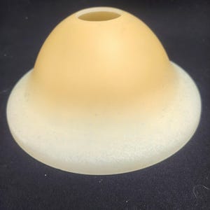 Puede incluir: Una pantalla de lámpara de vidrio en forma de cúpula con un acabado mate suave que transiciona de un color ámbar cálido y sólido en la parte superior a un tono crema más claro y moteado hacia el borde inferior ancho, con una abertura circular.