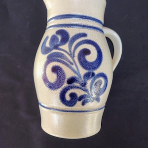 Pode incluir: Uma jarra de grés bege com textura salpicada e uma alça curva. O corpo é decorado com um padrão floral de volutas pintado à mão em azul escuro, acentuado por duas faixas azuis horizontais que circundam o pescoço e a seção inferior.