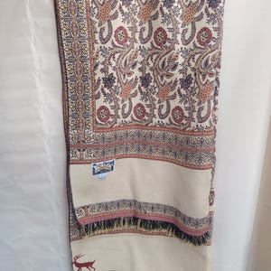 Pode incluir: Um xale retangular de cor creme com um padrão denso e intrincado de paisley e flores em tons de vermelho, castanho e azul escuro. O tecido inclui uma borda tecida, uma pequena etiqueta retangular da marca e uma silhueta de veado vermelho.