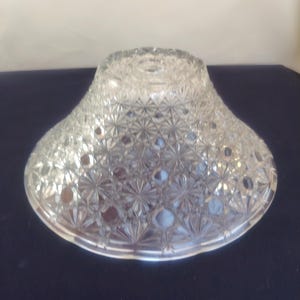 May include: A clear glass bowl featuring an intricate pressed pattern of repeating starbursts and geometric shapes. The bowl has a wide, flared rim and a circular base, with a reflective surface that catches light across its textured exterior.