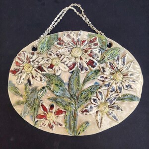 Op de afbeelding: Een ovaal keramieken wandbord met een reliëfontwerp van witte madeliefjes met donkerrode en blauwe accenten, groene bladeren en gele harten. Het stuk heeft een glanzende afwerking en bevat een zilverkleurige metalen ketting om op te hangen.
