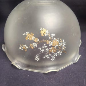 Op de afbeelding: Een koepelvormige lampenkap van matglas met een geschulpte rand en een centraal bloemmotief. Het patroon bestaat uit fijne takken met kleine goudkleurige bloesems en witte bladeren, wat zorgt voor een gedetailleerd botanisch ontwerp op het oppervlak.