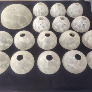 Könnte beinhalten: Eine Sammlung von sechzehn kuppelförmigen Lampenschirmen aus Glas mit einer durchscheinenden, marmorierten weißen und cremeweißen Oberfläche. Jeder Schirm hat eine kreisförmige Öffnung an der Oberseite zur Montage, mit einem größeren Schirm oben links.