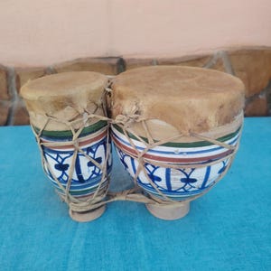 Peut inclure: Une paire de bongos reliés avec des corps en céramique peints à la main présentant des motifs géométriques bleus, verts et marron. Les tambours ont des peaux naturelles couleur beige fixées par un réseau de cordes claires sur l'extérieur.