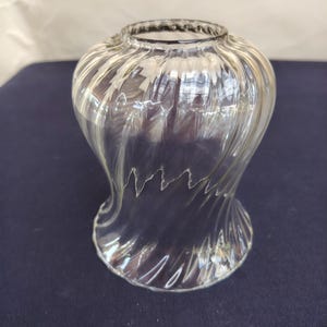 Könnte beinhalten: Ein Lampenschirm aus klarem Glas mit einer glockenförmigen Silhouette und einem ausgeprägten spiralförmigen Rippenmuster. Das Glas ist transparent und glatt, mit einer kreisförmigen Öffnung oben und einer breiteren, ausgestellten Öffnung.