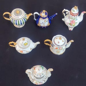 Può includere: Una collezione di sei teiere in miniatura in porcellana con vari design, tra cui strisce blu e verdi, un corpo blu intenso con illustrazioni di uccelli e diverse teiere bianche decorate con motivi floreali colorati e dettagli dorati.